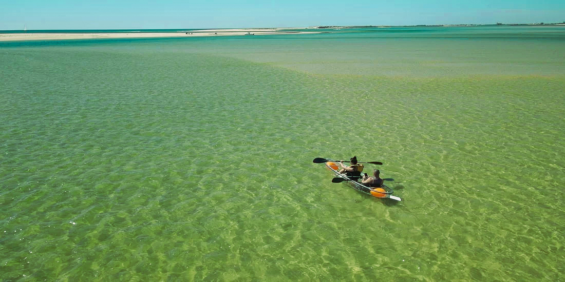 Two people in a transparent kayak on shallow green water, with a sandbank on the horizon.